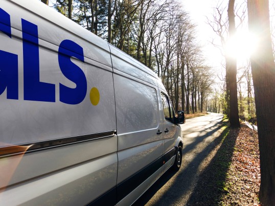 GLS delivery van driving on a tree-lined road, illuminated by sunlight.