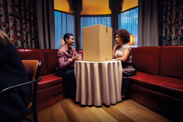 Deux personnes dînent dans un restaurant, séparées par un grand carton posé sur la table, illustrant la rupture dans l’expérience client. Une métaphore visuelle utilisée par GLS pour souligner l’importance d’une livraison fluide et personnalisée, enjeu clé présenté lors du salon Tech For Retail 2025.