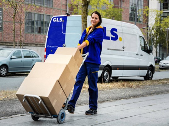 GLS delivery woman transporting multiple parcels on a hand truck, with a GLS delivery van in the background.